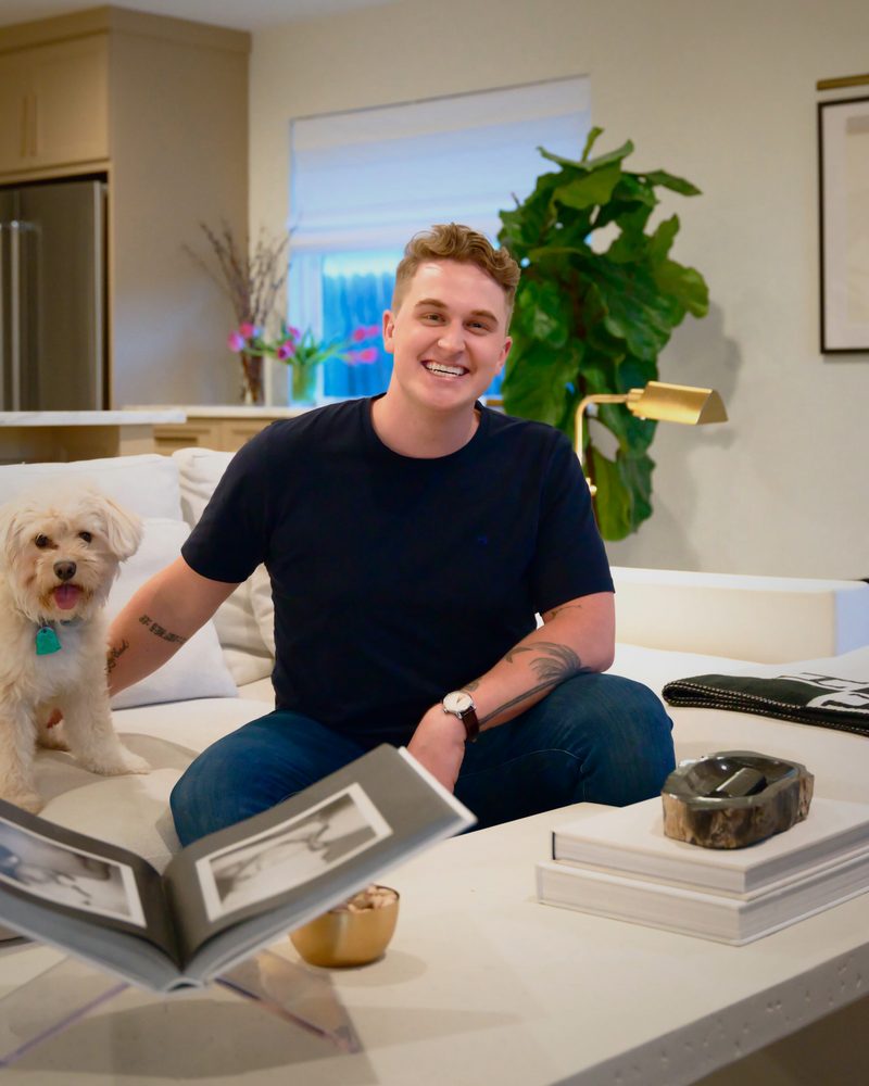 Spenser Marriot on a white sofa beside a small white dog, an open book on the coffee table in front of him