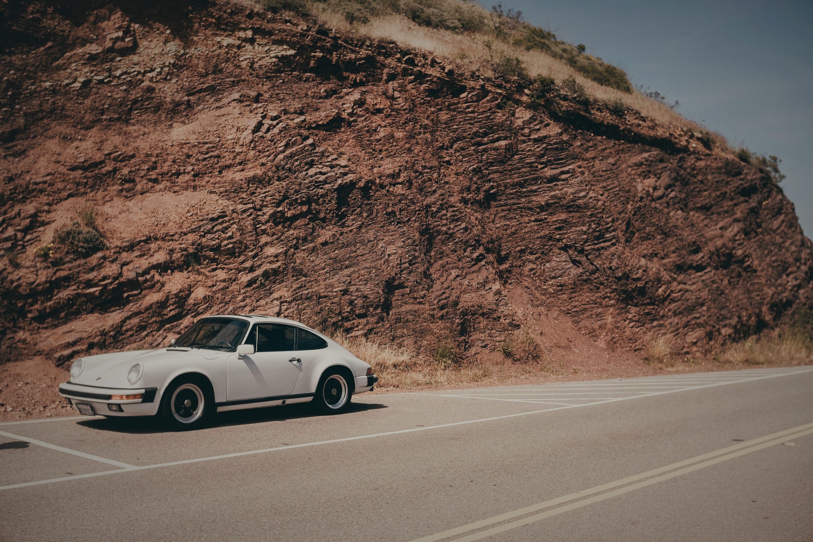 A white vintage Porsche 911 on a coastal road under a red rock cliff