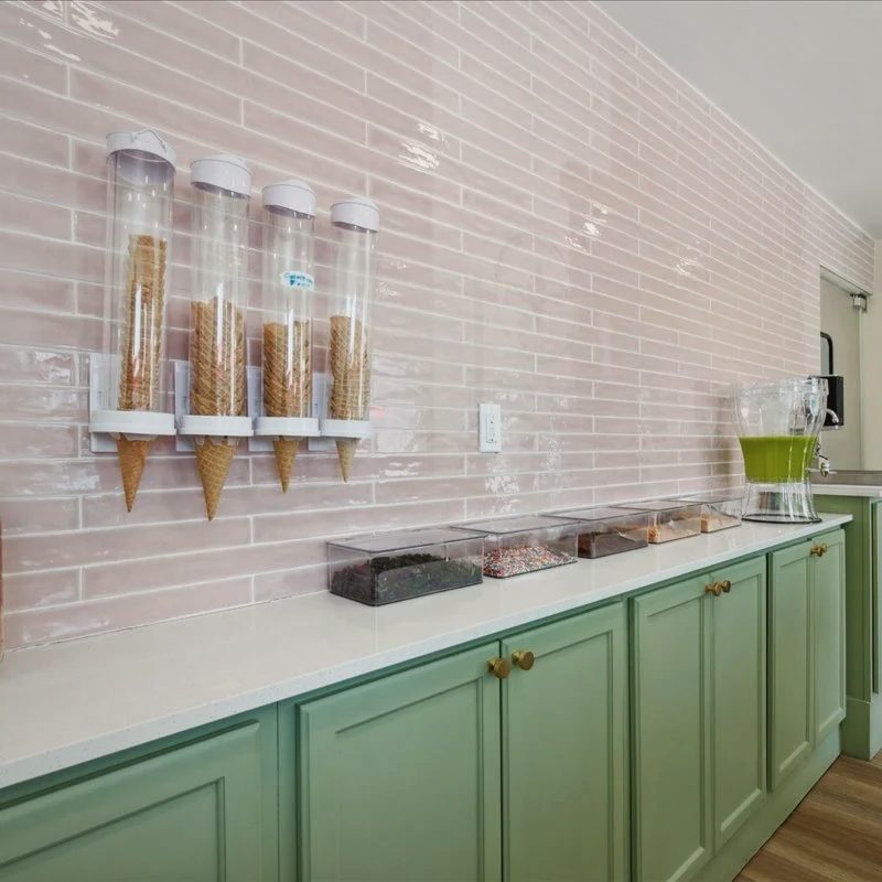 Ice cream shop counter with green cabinets and brass pulls under a pink tile wall, waffle cones in glass dispensers