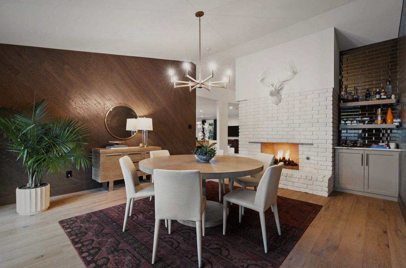 Dining room with a round table and white chairs on a dark rug, a brass chandelier, a wood-paneled wall and a white brick fireplace