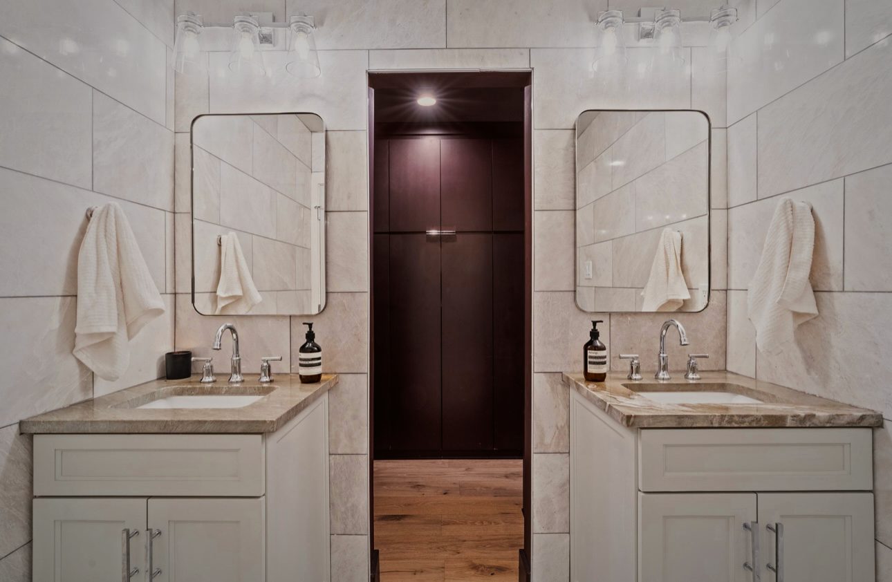 Primary bath with two white vanities, marble walls, round mirrors and a dark doorway between them