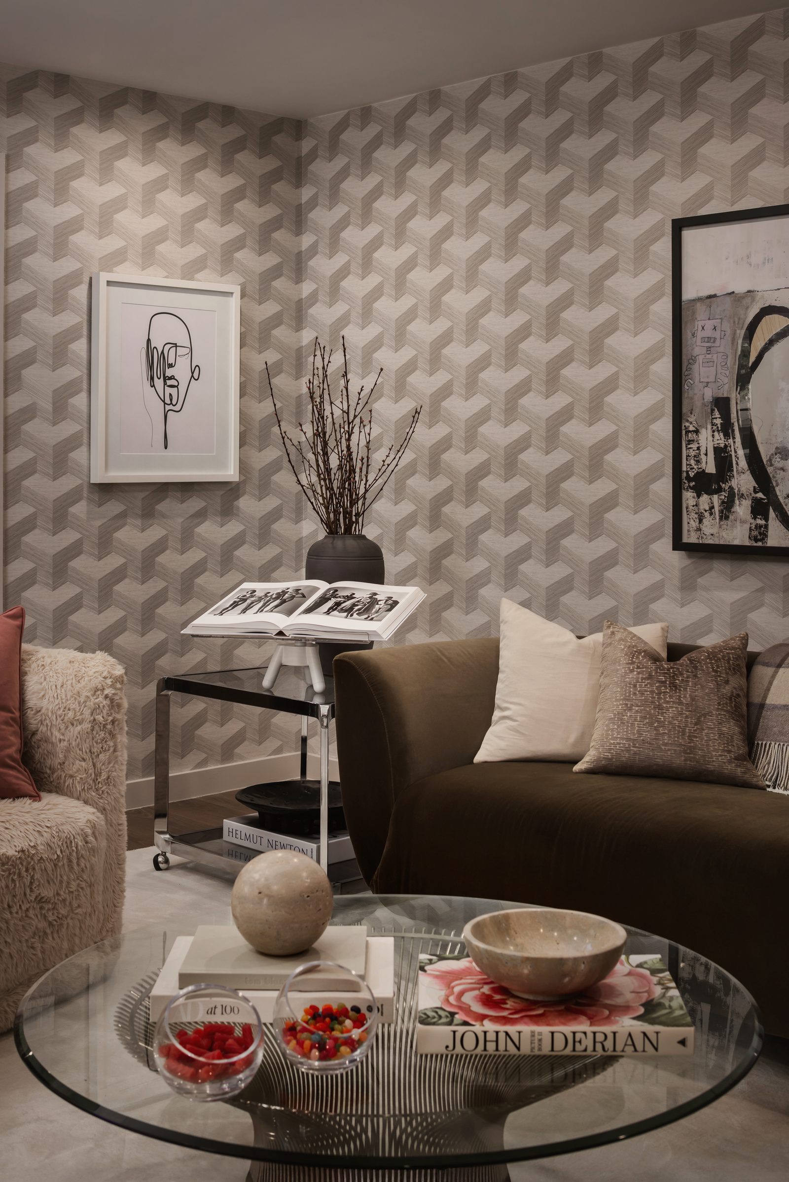 Den with gray geometric wallpaper, a brown velvet sofa, a glass coffee table and framed black-and-white art