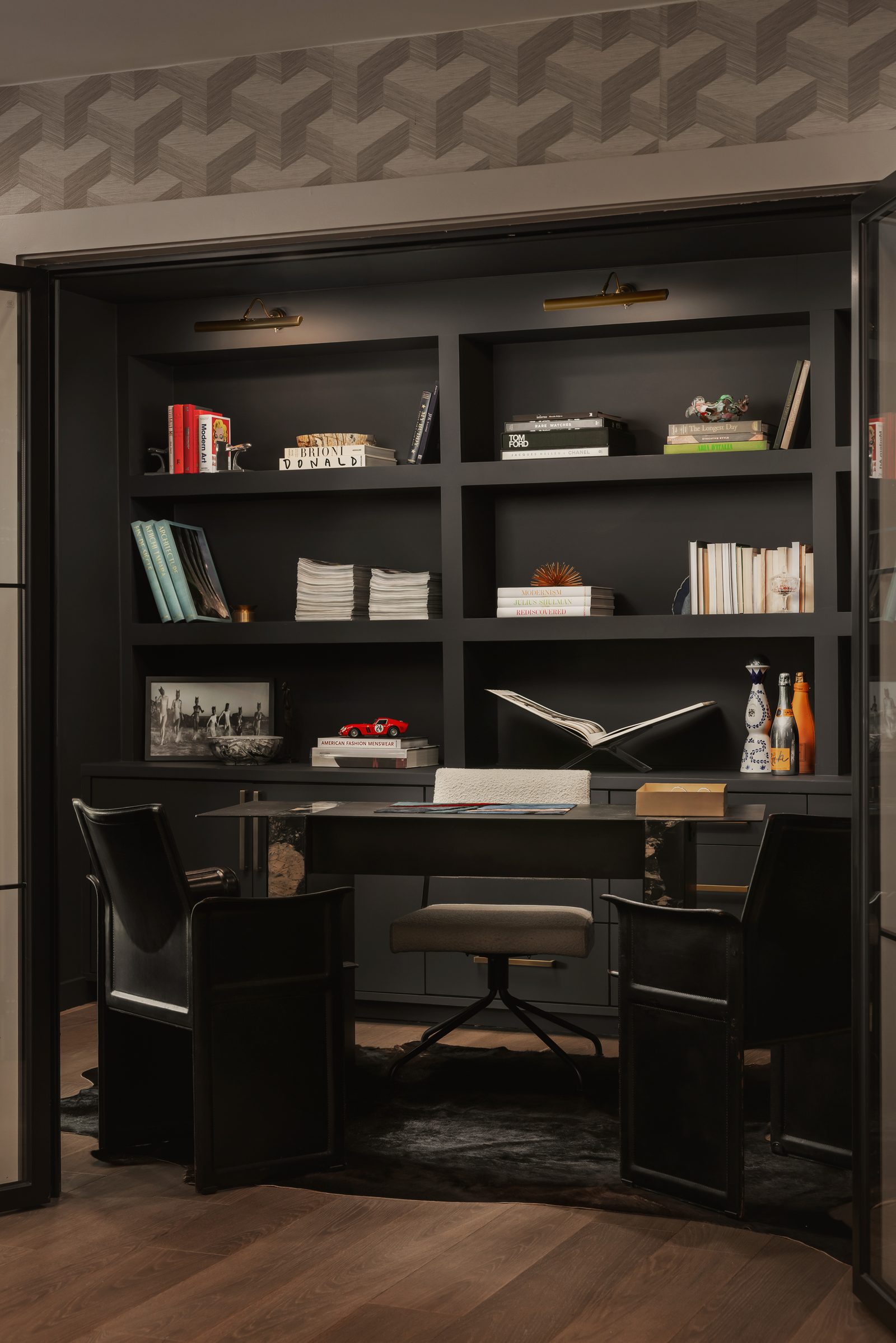 Black built-in desk and shelves set into a wall, with a black chair and brass picture lights