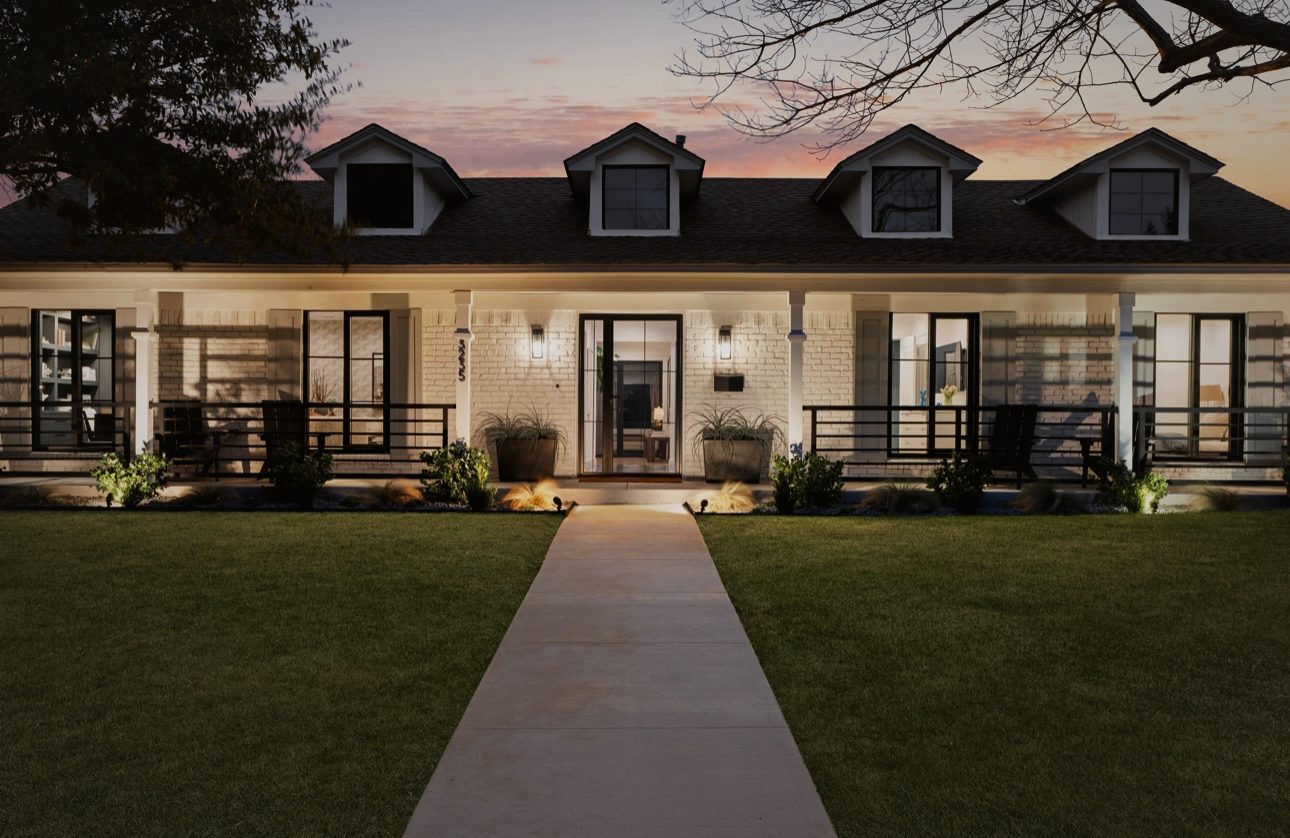 The house at dusk: a white brick front with three dormers, a long porch with black railings and a walk across the lawn