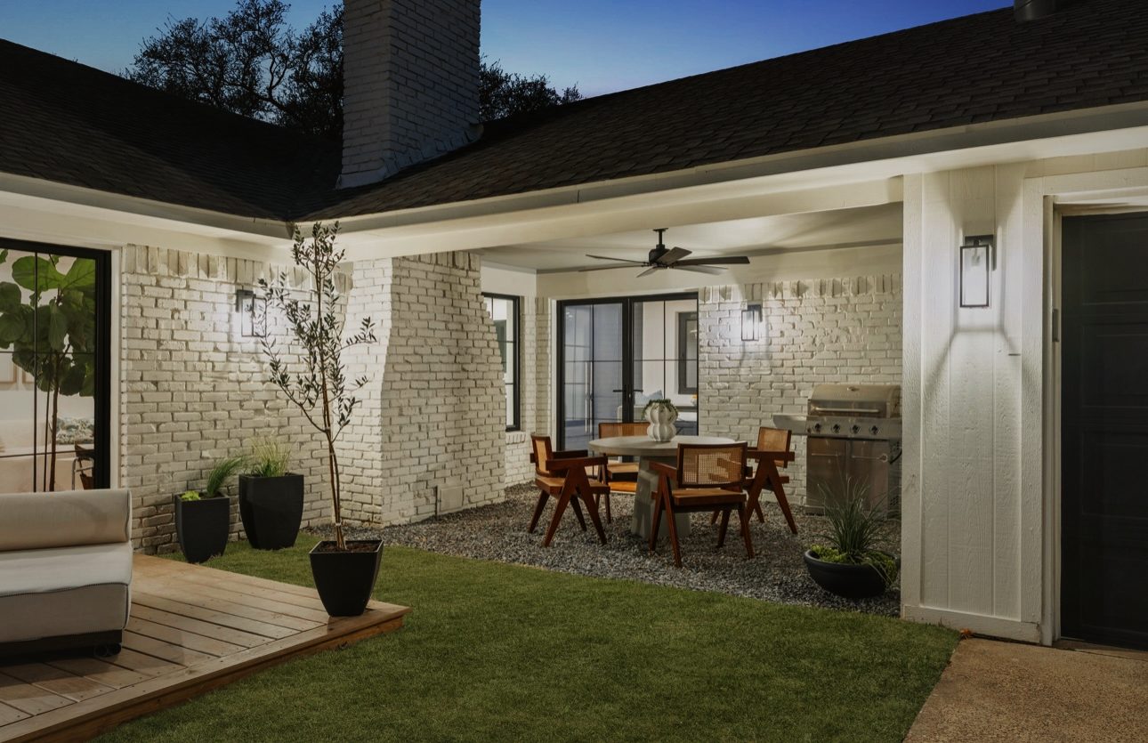 Covered patio at dusk with a round dining table, wood chairs, a grill and white brick walls