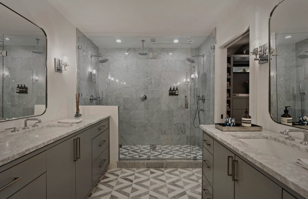 Primary bath with gray vanities, marble counters, a glass shower and a patterned marble floor