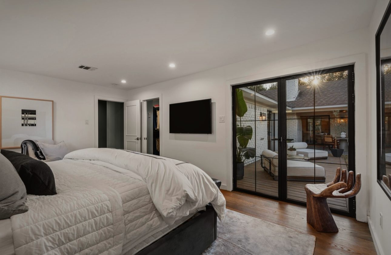 Primary bedroom with a white bed, black steel doors onto the patio and a carved wood chair