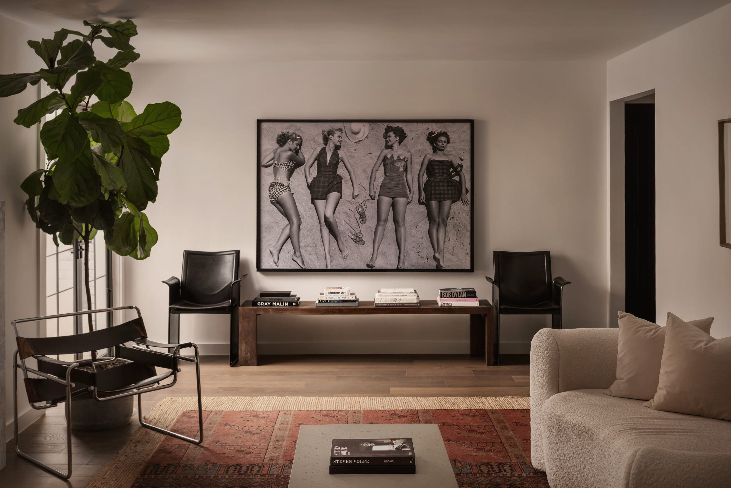 Sitting room with a large black-and-white beach photograph over a walnut bench, black leather sling chairs and a fiddle-leaf fig